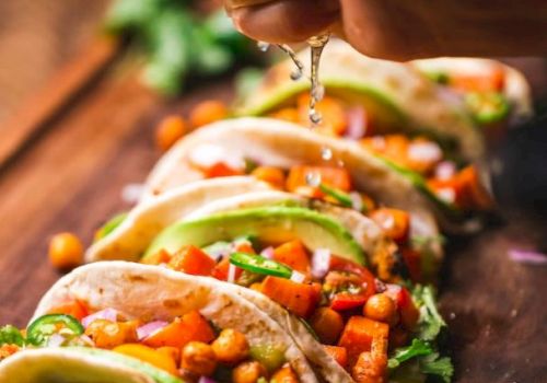 Hand squeezing lime over tacos filled with chickpeas, avocado slices, diced vegetables, and garnished with cilantro on a wooden board.