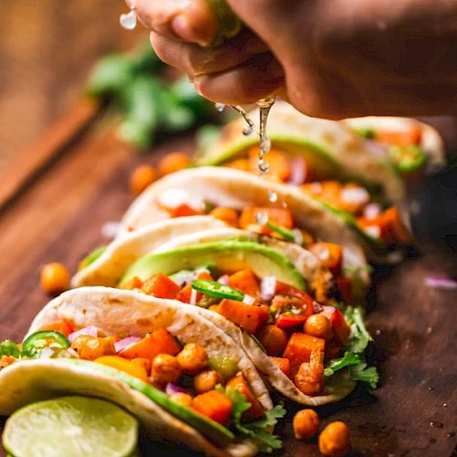 Hand squeezing lime over tacos filled with chickpeas, avocado slices, diced vegetables, and garnished with cilantro on a wooden board.