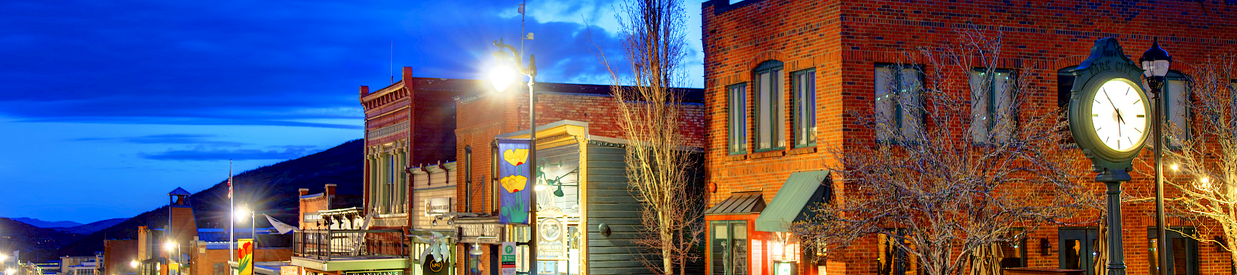 A quiet small-town street at dusk, brick storefronts, warm lights, clock on a post, empty sidewalk and blue evening sky.