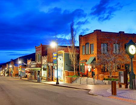 A quiet small-town street at dusk, brick storefronts, warm lights, clock on a post, empty sidewalk and blue evening sky.