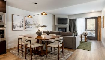 A modern open-concept living and dining area with a wood dining table and six chairs, a gray sectional, a TV above a fireplace, framed wall art, and large windows letting in natural light.