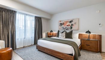 A modern hotel room with a wooden bed, two matching nightstands, gray bedding, decorative pillows, a large window with dark curtains, and wall art.