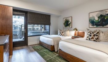 A bright hotel room with two queen beds, a wooden dresser, and a large window/door leading to a balcony; modern decor with framed art and a green rug.