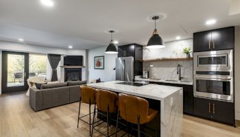 A modern open-plan kitchen and living area with an island, pendant lights, sofa, and built-in ovens, featuring a sleek and cozy design.