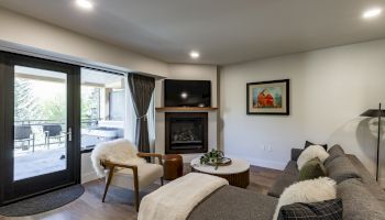 A cozy living room with a sectional sofa, fireplace, wall art, and a view of the outdoor patio through glass doors.