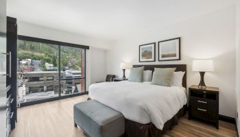 A modern bedroom with a large bed, two lamps, framed art, and a window view of a cityscape and greenery adorns the space.