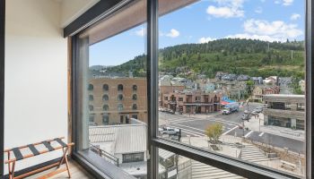 A view from a modern window shows a bustling street, historic buildings, and green hills under a blue sky with clouds.