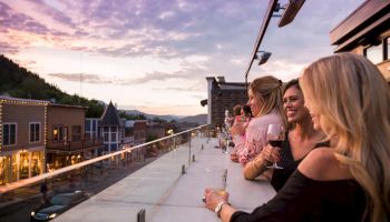 A group of friends on a rooftop terrace at sunset, chatting and enjoying drinks with a scenic town view in the background.