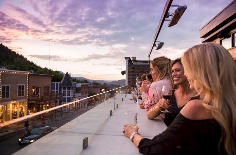 A group of friends on a rooftop terrace at sunset, chatting and enjoying drinks with a scenic town view in the background.
