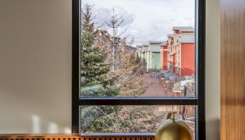 A view through a tall window shows a tree, a row of colorful buildings outside, with a wooden radiator shelf and a brass desk lamp inside, bathed in light.