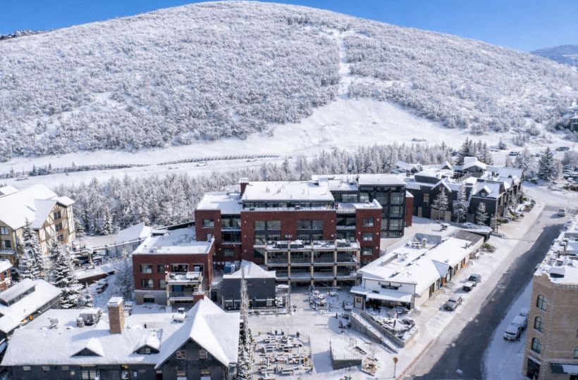 Snow-covered mountain village with modern buildings, cars, and a winding snow trail on the slope, under a clear blue sky.