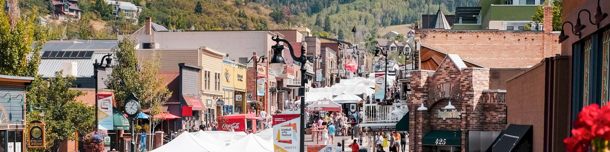 A bustling street fair with tents and people, surrounded by buildings and greenery, set against a backdrop of forested hills.