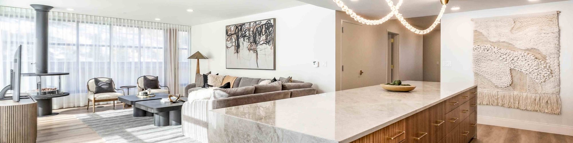 Bright modern living room/kitchen with gray sectional, island bar, gold chandelier lights, wood cabinets, and wall art decor.