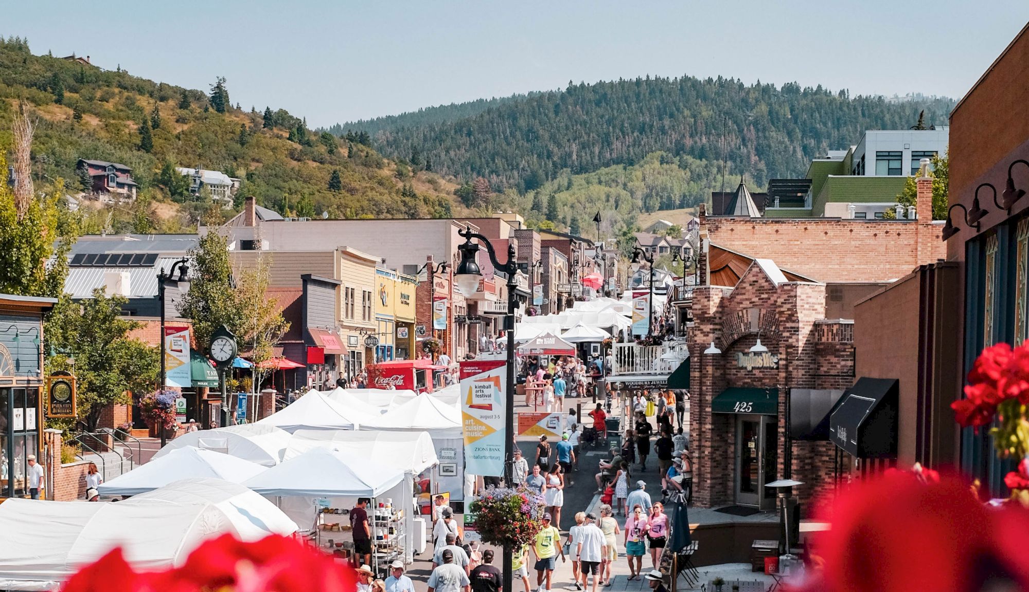 A lively street scene with people, tents, and shops set against a backdrop of green hills and a clear sky.