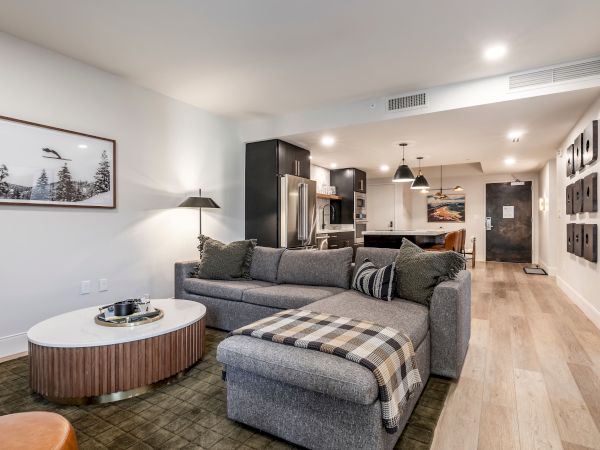 Modern open-plan living room with a gray sectional sofa, coffee table, framed art, and stainless kitchen appliances in view.