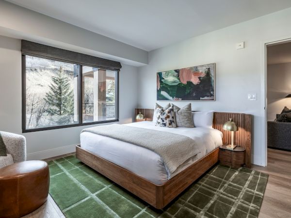 A modern bedroom with a large bed, window with blinds, abstract wall art, and warm wood nightstands and lamps.