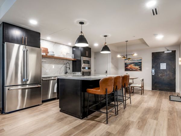 A modern kitchen with stainless steel appliances, a dark island with bar stools, pendant lights, and a marble-style backsplash.