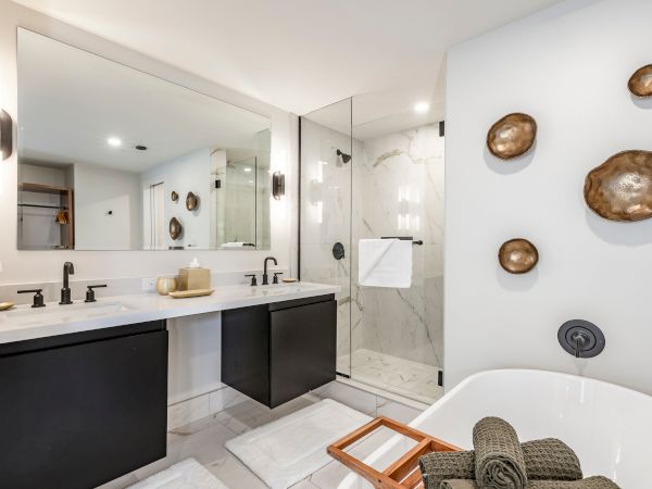 Modern bathroom with a vanity, glass shower, marble tile, towel, bronze wall decor, and a bathtub in the foreground.