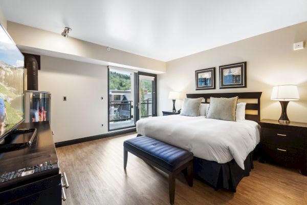 A modern hotel room featuring a large bed, seating area, TV, lamps, and a window with a view, decorated in neutral tones.