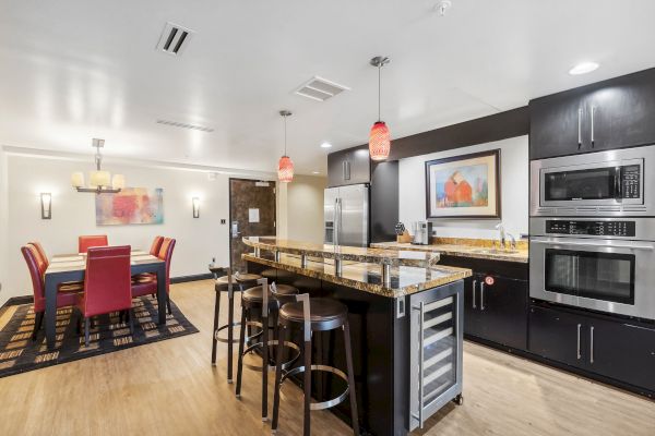 Modern kitchen and dining area with granite countertops, bar stools, red dining chairs, stainless steel appliances, and artwork on the wall.