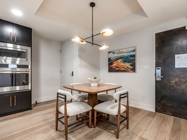 A modern dining area with a wooden table and chairs, pendant lights, a kitchen appliance wall, and framed artwork.