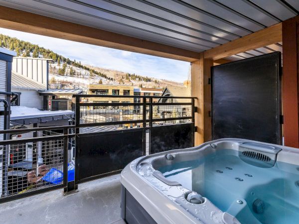 A covered balcony with a hot tub beside a snowy mountain view, framed by wooden beams and a fence.