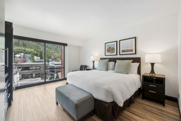 A modern bedroom with a large bed, two nightstands, lamps, artwork, and a balcony view outside the window.