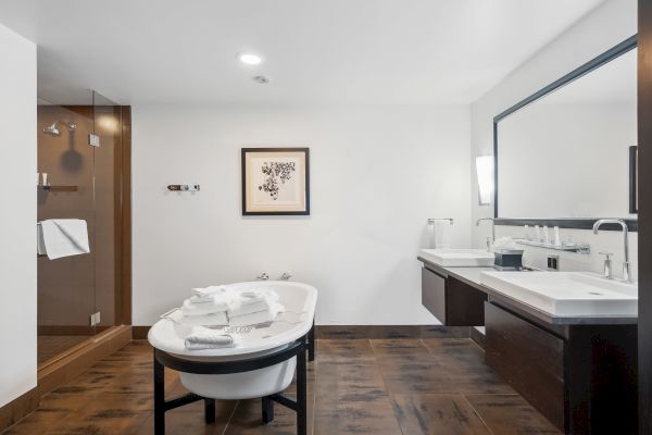 A modern bathroom with a white bathtub, wooden floor, large mirror, dual sinks, and a glass shower.