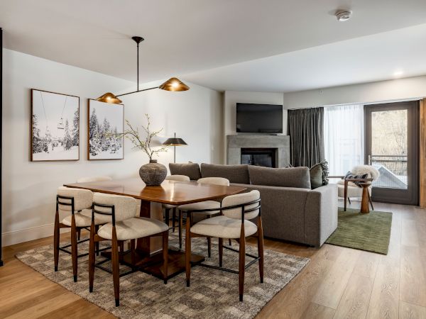 Bright living room with a dining table, sofas, TV above a fireplace, pendant lights, and winter forest wall art.