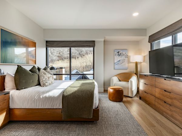 A modern bedroom with a bed by large windows, framed art, a dresser with TV, and warm neutral decor.