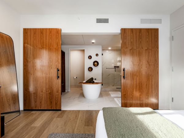 Modern bathroom with open wooden doors, white soaking tub, marble shower, and towel on a grey bath mat.