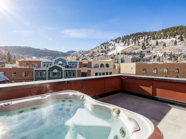 A rooftop hot tub overlooks a small town with brick buildings and snowy pine-covered hills under a bright blue sky.