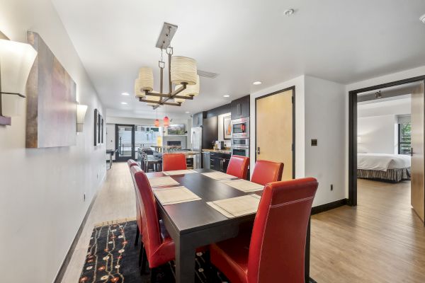 Modern dining area with a black table, red chairs, pendant lights, and an adjoining kitchen. Bedroom is visible through an open door.