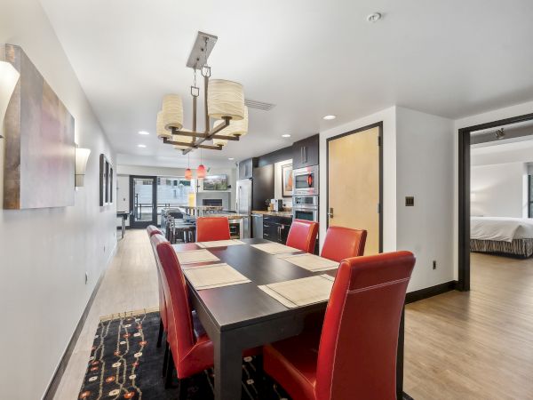 Modern dining area with a black table, red chairs, pendant lights, and an adjoining kitchen. Bedroom is visible through an open door.