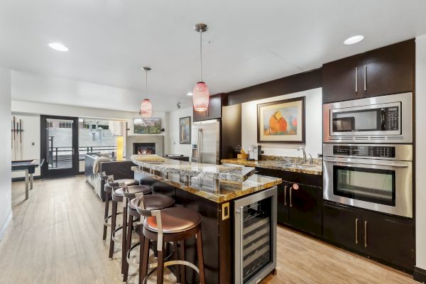 A modern kitchen with a bar counter, high stools, stainless steel appliances, pendant lights, and an open layout to a living area.