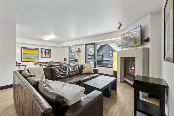 A cozy living room with a leather sofa, fireplace, wall-mounted TV, and pool table. Large windows offer a view of the outside.