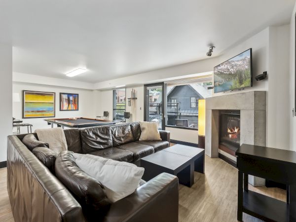 A cozy living room with a leather sofa, fireplace, wall-mounted TV, and pool table. Large windows offer a view of the outside.