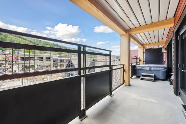 A covered balcony with a railing and a hot tub overlooks a scenic view of buildings and hills under a blue sky with clouds.