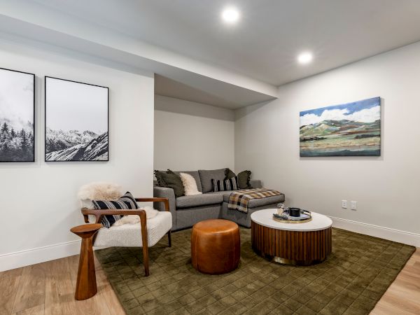 A modern living room with a gray sofa, wooden chair, round coffee table, ottoman, and framed landscape paintings under bright lights.