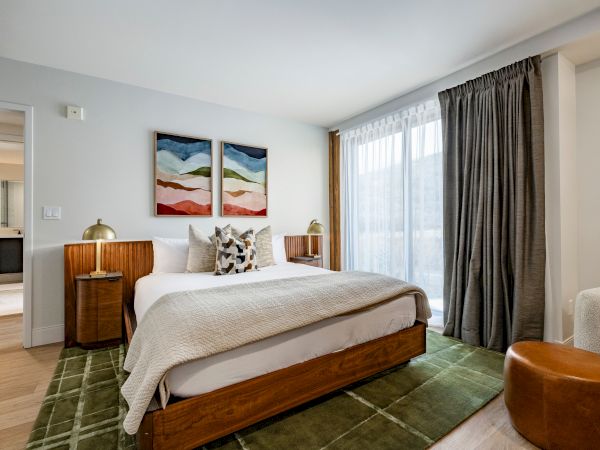 A bright bedroom with a wooden bed, white bedding, two landscape paintings, curtains, and bedside lamps.