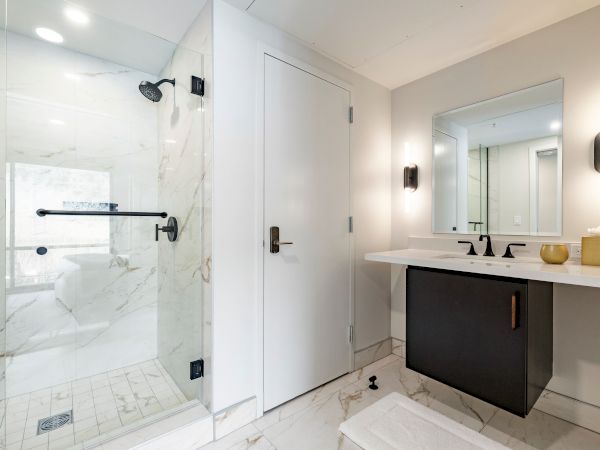 Modern bathroom with a glass shower, white marble walls, and a sleek vanity with black sink basins and round lights.