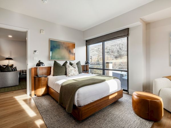 A modern bedroom with a wooden platform bed, pillows, large window with blinds, framed art, and warm sunlight.