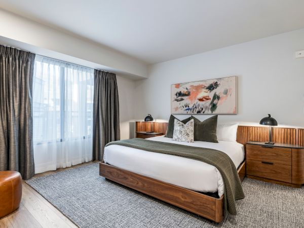 A modern bedroom with a wooden bed, gray curtains, bedside lamps, an abstract wall painting, and a patterned rug.