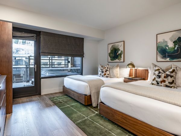 A modern hotel bedroom with two beds, patterned pillows, a green rug, and framed artwork by a window with blinds.