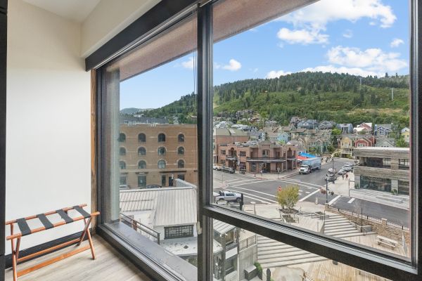 A room with large windows overlooks a scenic town street, surrounded by green hills under a blue sky with clouds.