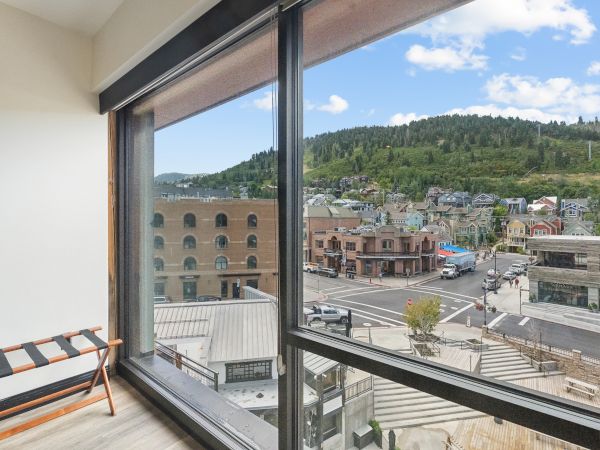 A room with large windows overlooks a scenic town street, surrounded by green hills under a blue sky with clouds.