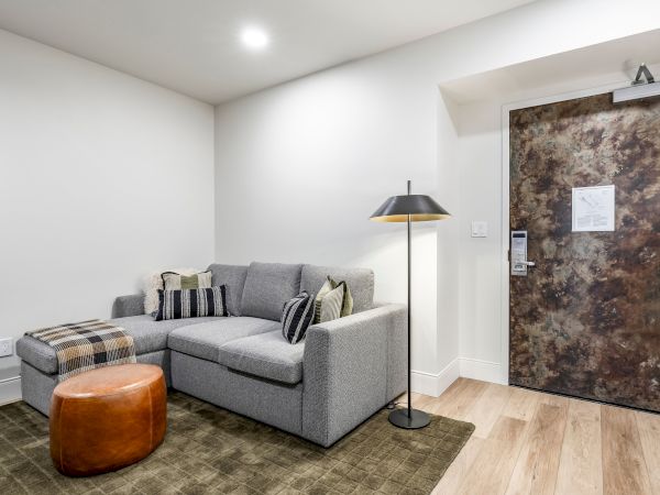 Bright lounge with a gray sectional sofa, striped pillows, floor lamp, ottoman, rug, and a brown decorative door with sign.