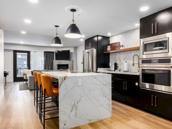 A modern kitchen with a marble island, dark cabinets, pendant lights, a stainless range, and an open dining/living area beyond.
