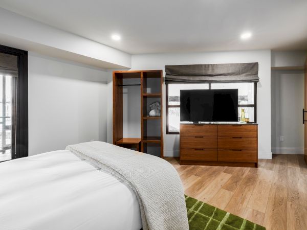 A modern bedroom with a bed, wooden TV console, built-in shelving, blinds by the window, and warm wood flooring.