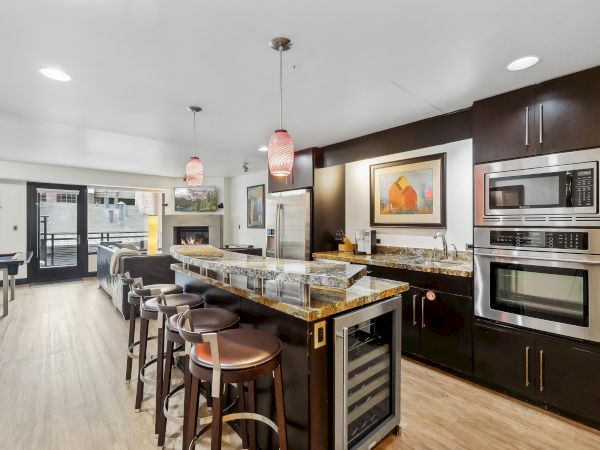 Modern kitchen with a bar, stools, stainless steel appliances, pendant lights, and a living area in the background.